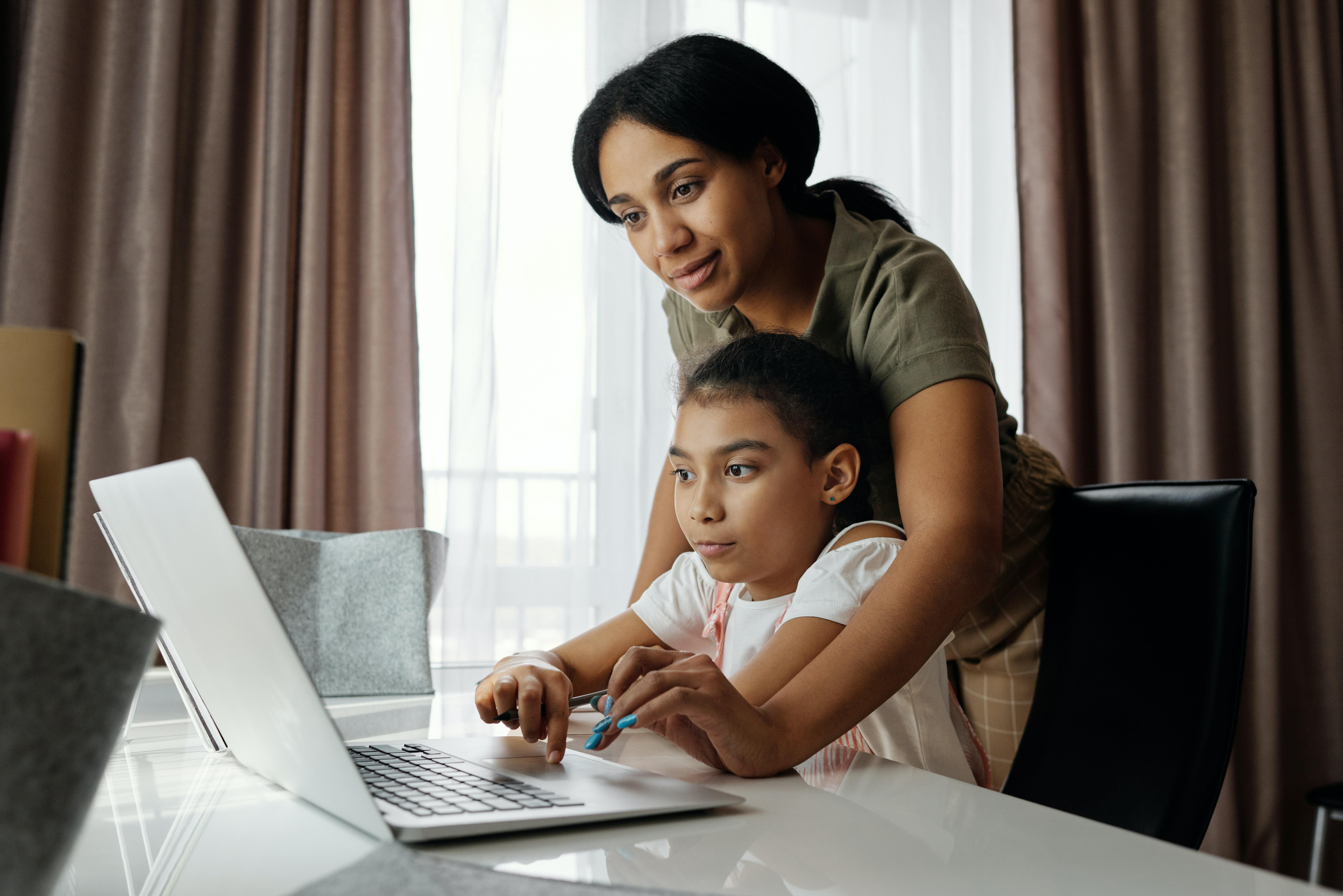 A woman standing over a child pressing a laptop