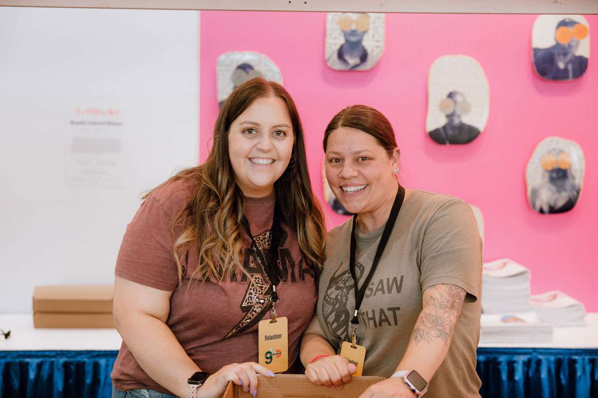 Two female convention volunteers wearing event badges smiling in front of a pink display wall