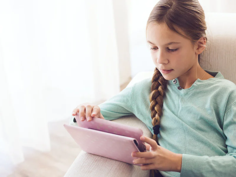 A young girl with a long braid wearing a light blue sweater, looking intently at a pink tablet