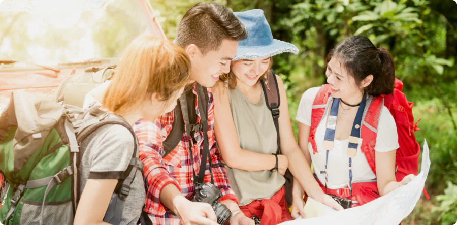 A mother, father, and two daughters wearing backpacks and smiling as they look at a large paper map together while hiking in a lush green forest