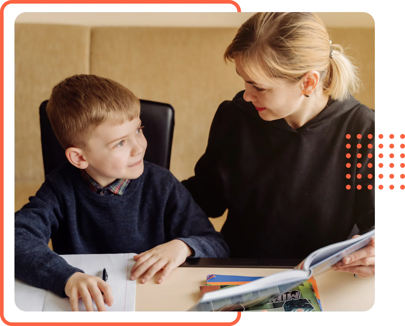 A young boy holding a pen at a desk while his mother sits beside him holding an open workbook during a homeschool lesson