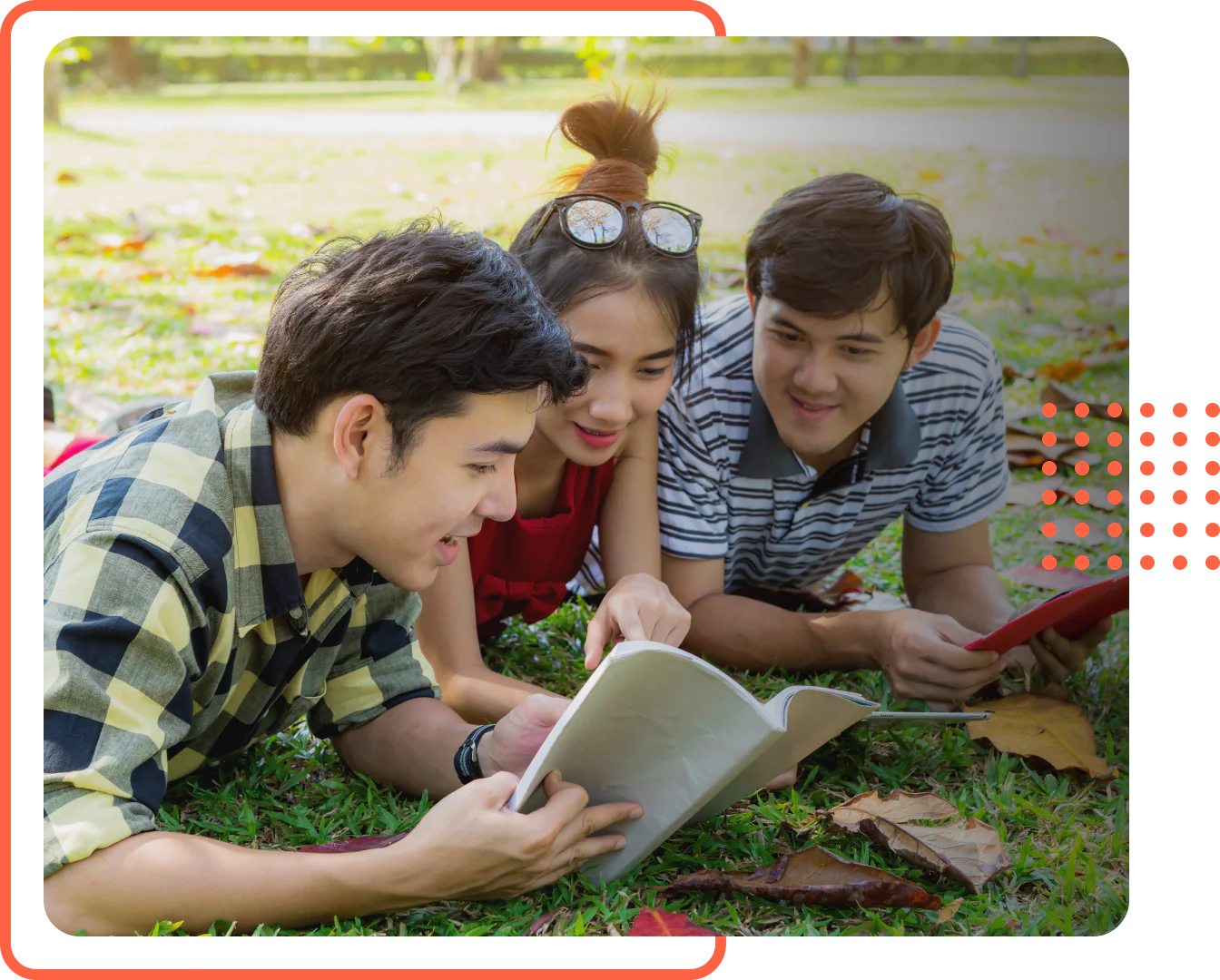 Three young adults lying on the grass outdoors, smiling and looking at an open book together while studying.