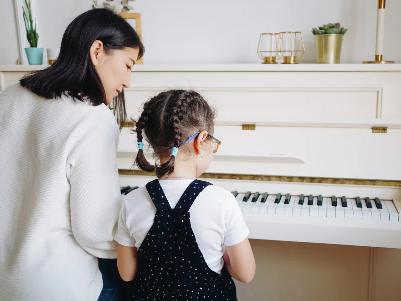 A woman and a young girl with braided hair sitting together at a white piano during a music lesson