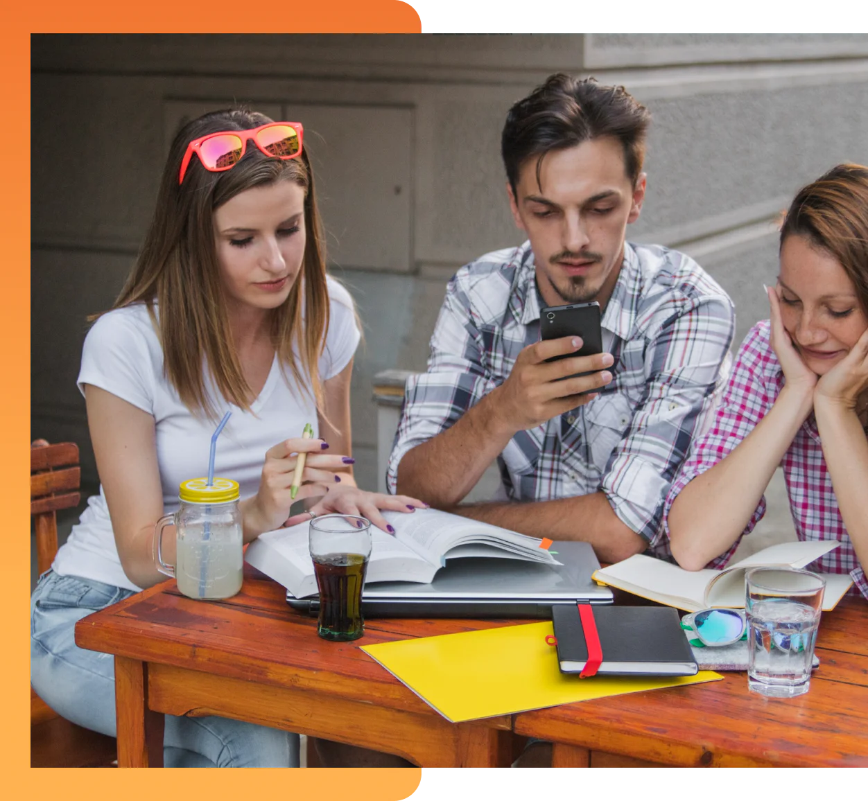 Three college-aged students sitting around a wooden outdoor table looking at open books, notebooks, and a smartphone
