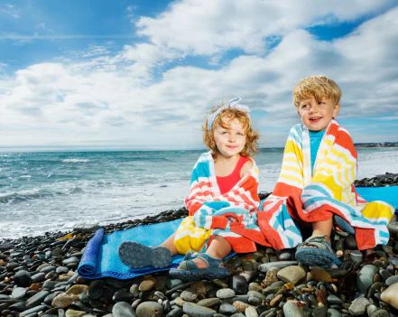 A young boy and girl sitting on a rocky beach wrapped in bright striped towels with the ocean in the background