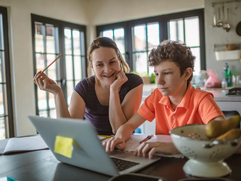 A mother holding a pencil and smiling while her teenage son works on a laptop at a kitchen counter