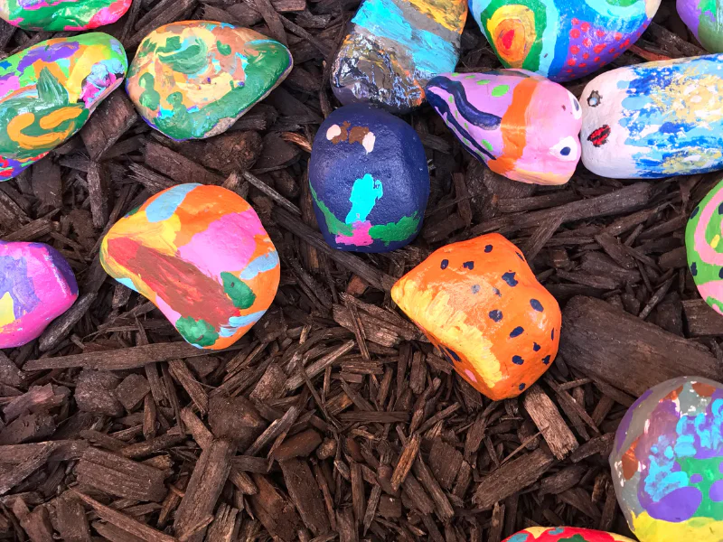 A collection of brightly painted rocks with various patterns and designs resting on dark wood chips during a hands-on learning activity