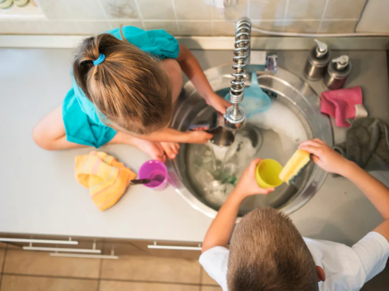 An overhead view of a young boy and girl washing a cup and a bowl with sponges in a soapy kitchen sink