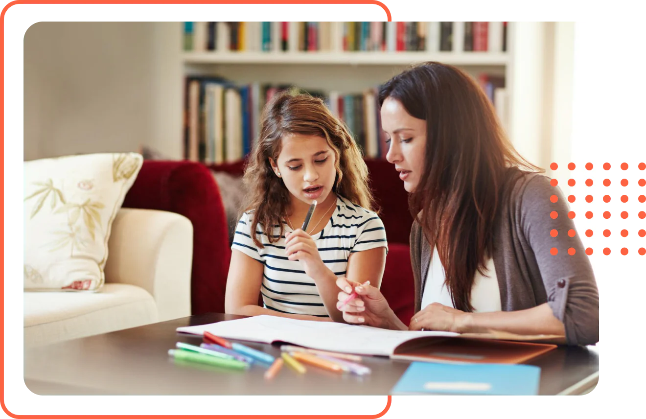 A mother and her young daughter sitting in a living room looking at an open workbook with colored pencils during a homeschool lesson