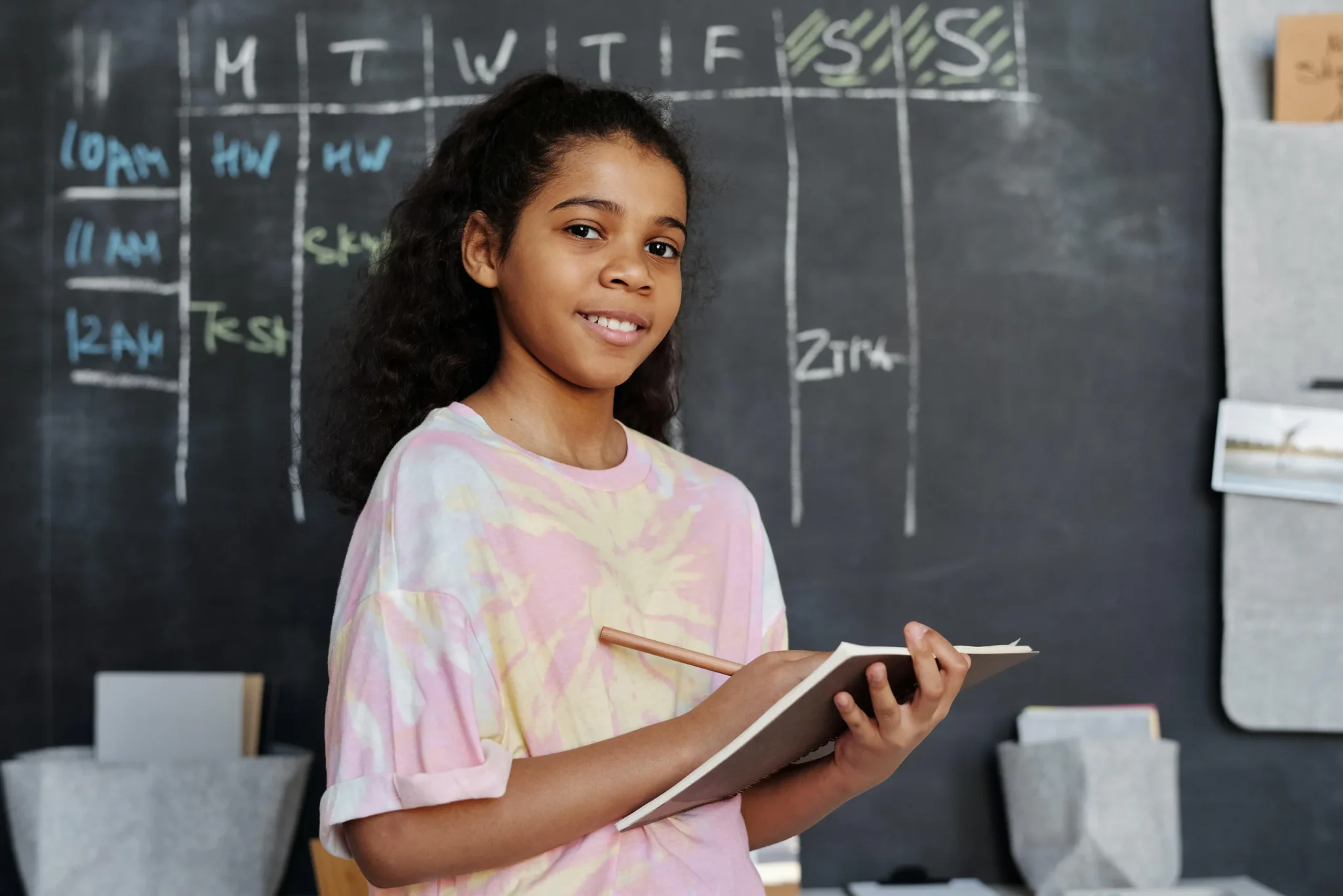 A child holding a pen and a book, standing in front of a black chalkboard