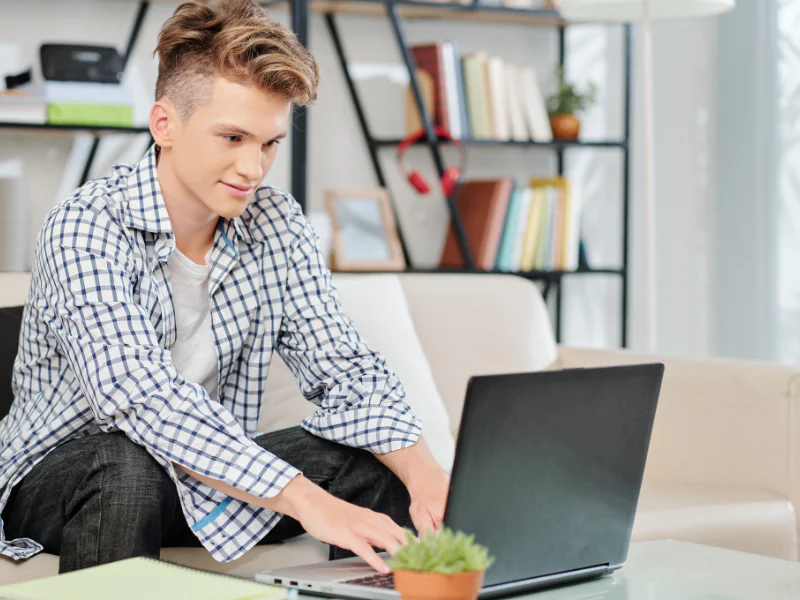 A teenage high school student sitting by a couch and typing on a laptop during a homeschool lesson