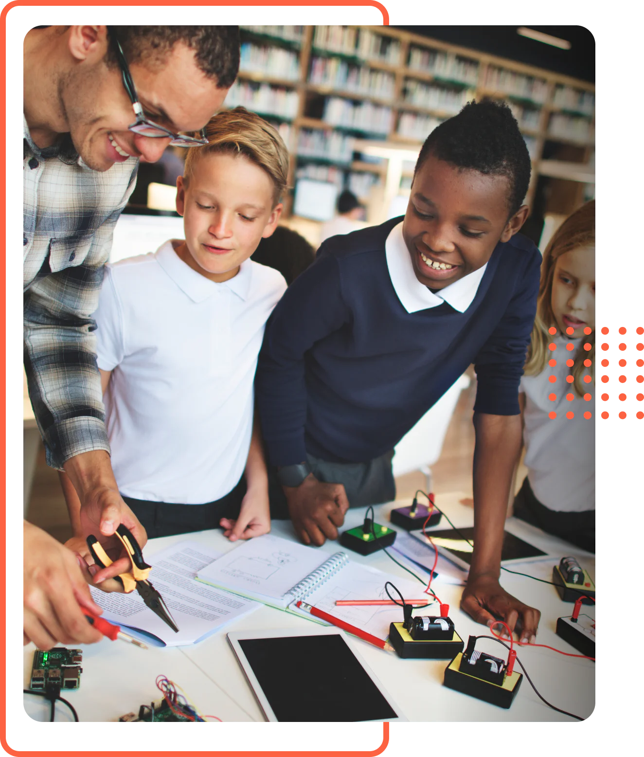 A smiling male teacher helping three young students build an electrical circuit at a desk