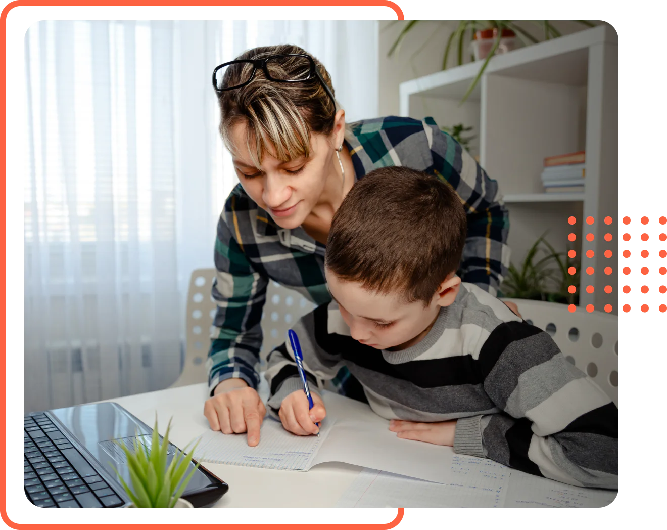 A woman and a young boy looking at a notebook and writing at a white desk in a home setting